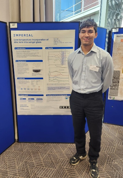 Photograph of a person with short black hair wearing a light blue shirt, black trousers and black trainers stood in front of a poster